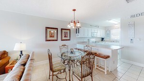 A modern kitchen with a central island, white cabinetry, and a dining area with a glass table and cushioned chairs. - Perfectly located condo in beachfront complex with pool, sauna & WiFi (Orange Beach)