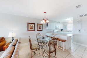 A modern kitchen with a central island, white cabinetry, and a dining area with a glass table and cushioned chairs. - Perfectly located condo in beachfront complex with pool, sauna & WiFi (Orange Beach)
