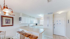 A kitchen with white cabinets, a central island with stools, a hanging light fixture, and a door with a sign. - Perfectly located condo in beachfront complex with pool, sauna & WiFi (Orange Beach)