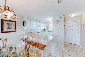 A kitchen with white cabinets, a central island with stools, a hanging light fixture, and a door with a sign. - Perfectly located condo in beachfront complex with pool, sauna & WiFi (Orange Beach)