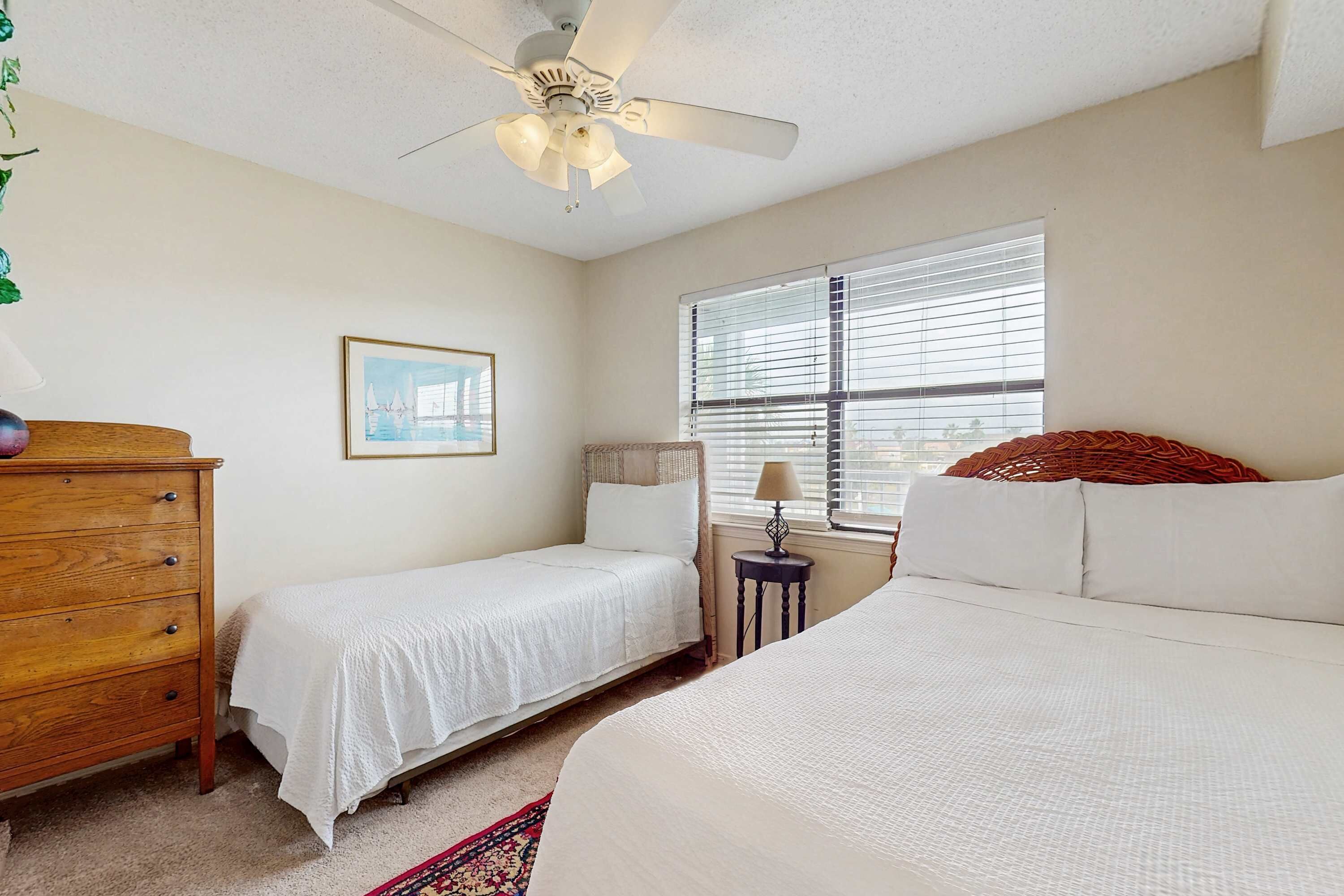 A bedroom with two beds, a wooden dresser, a ceiling fan, a window with blinds, and a framed picture on the wall.