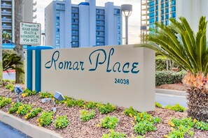A sign for 'Romar Place' with a number '24038' and a backdrop of modern buildings and a well-maintained garden. - Gulf-front condo with pool, hot tub, sauna, gym, AC, cable, & washer/dryer (Orange Beach)