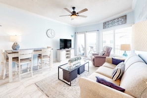 A living room with a ceiling fan, a TV, a sofa, a coffee table, and a dining area with chairs and a table. - Condo in beachfront complex with partial Gulf views, balcony, pools, & gym (Orange Beach)