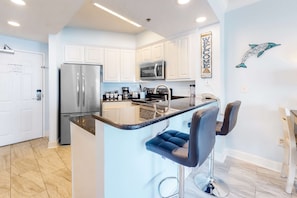 A modern kitchen with white cabinets, a stainless steel refrigerator, a microwave, and a bar area with two stools. - Condo in beachfront complex with partial Gulf views, balcony, pools, & gym (Orange Beach)