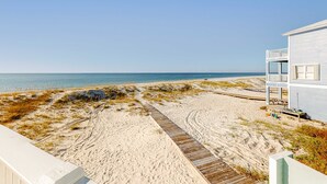 Vista de la playa con un paseo de madera que lleva a un edificio blanco con balcón.