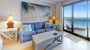 Una sala de estar con sofá, dos mesitas auxiliares, una mesa de centro, un cuadro y vista al mar a través de amplios ventanales.