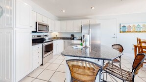 A kitchen with white cabinets, stainless steel appliances, a granite countertop, wicker chairs, and a painting of beach huts. - Gulf-front home on the beach with 3-tier deck, Gulf views & pool access (Orange Beach)