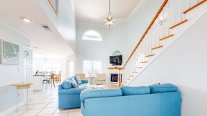 A spacious living room with a blue sofa, a dining area, a staircase, and a TV mounted on the wall. - Gulf-front home on the beach with 3-tier deck, Gulf views & pool access (Orange Beach)