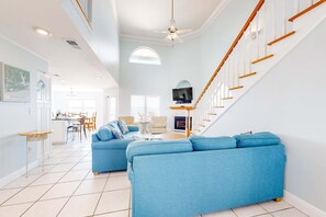 A spacious living room with a blue sofa, a dining area, a staircase, and a TV mounted on the wall. - Gulf-front home on the beach with 3-tier deck, Gulf views & pool access (Orange Beach)