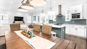 A modern kitchen with white cabinets, a blue backsplash, and stainless steel appliances. There is a dining area with a wooden table and patterned chairs. - Azure! Sleeps 22 ! Golf Cart ! Beach Access ! Pool! (Destin)