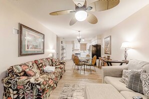 Ein Wohnzimmer mit Sofa, Couchtisch, Essbereich mit Stühlen, Deckenventilator und einer Küche im Hintergrund.
