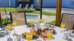 Une table dressée avec des aliments et des boissons pour le petit-déjeuner, donnant sur une piscine et un jardin.