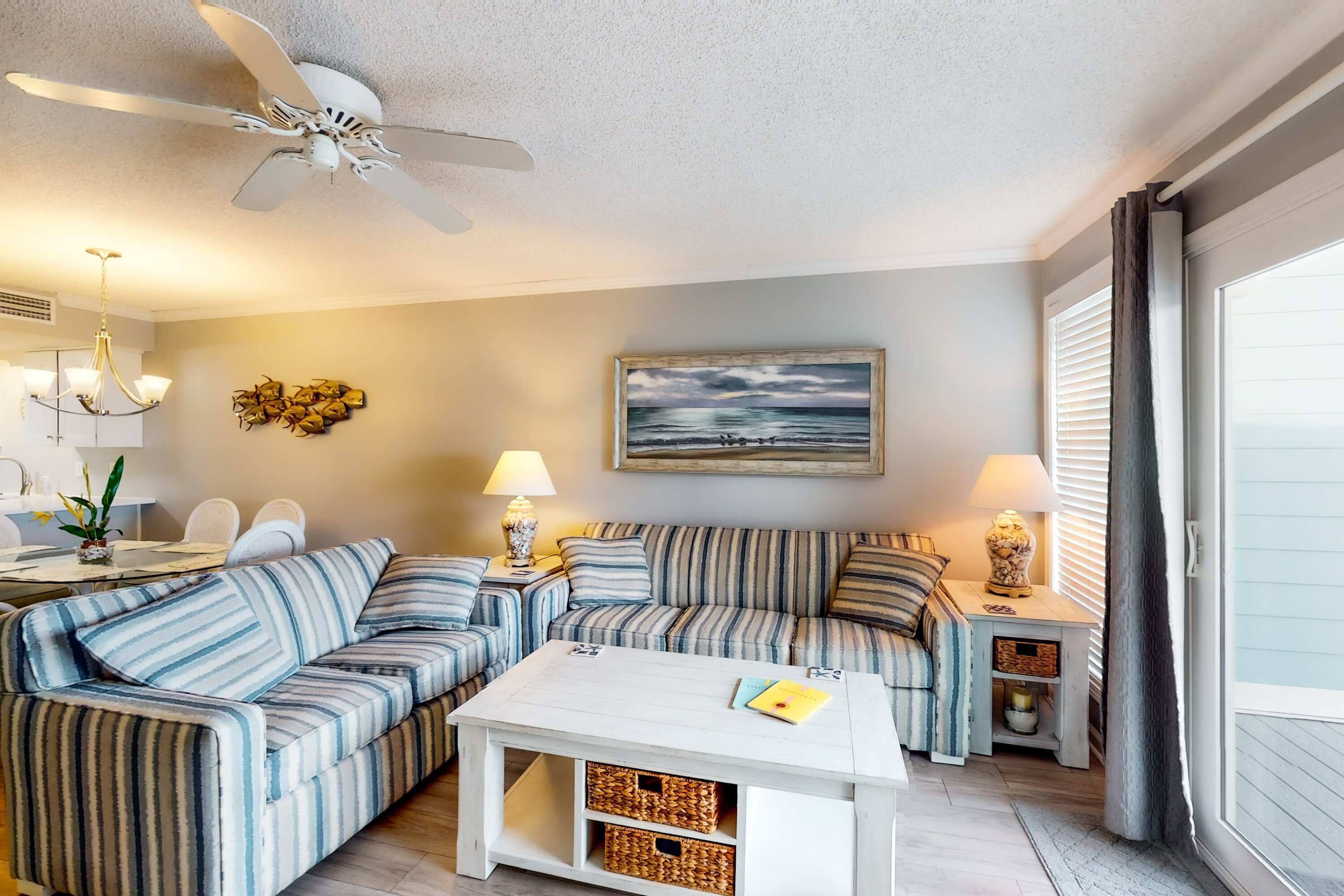 A living room with striped sofas, a white coffee table, a ceiling fan, a painting, and a dining area with a table and chairs.