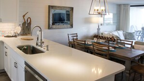 A modern kitchen with a central island, a dining area with wooden furniture, and a large window with curtains. - Coastal escape with 2 water-view balconies, 2 pools, tennis & beach access (Orange Beach)