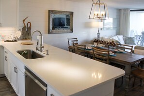 A modern kitchen with a central island, a dining area with wooden furniture, and a large window with curtains. - Coastal escape with 2 water-view balconies, 2 pools, tennis & beach access (Orange Beach)