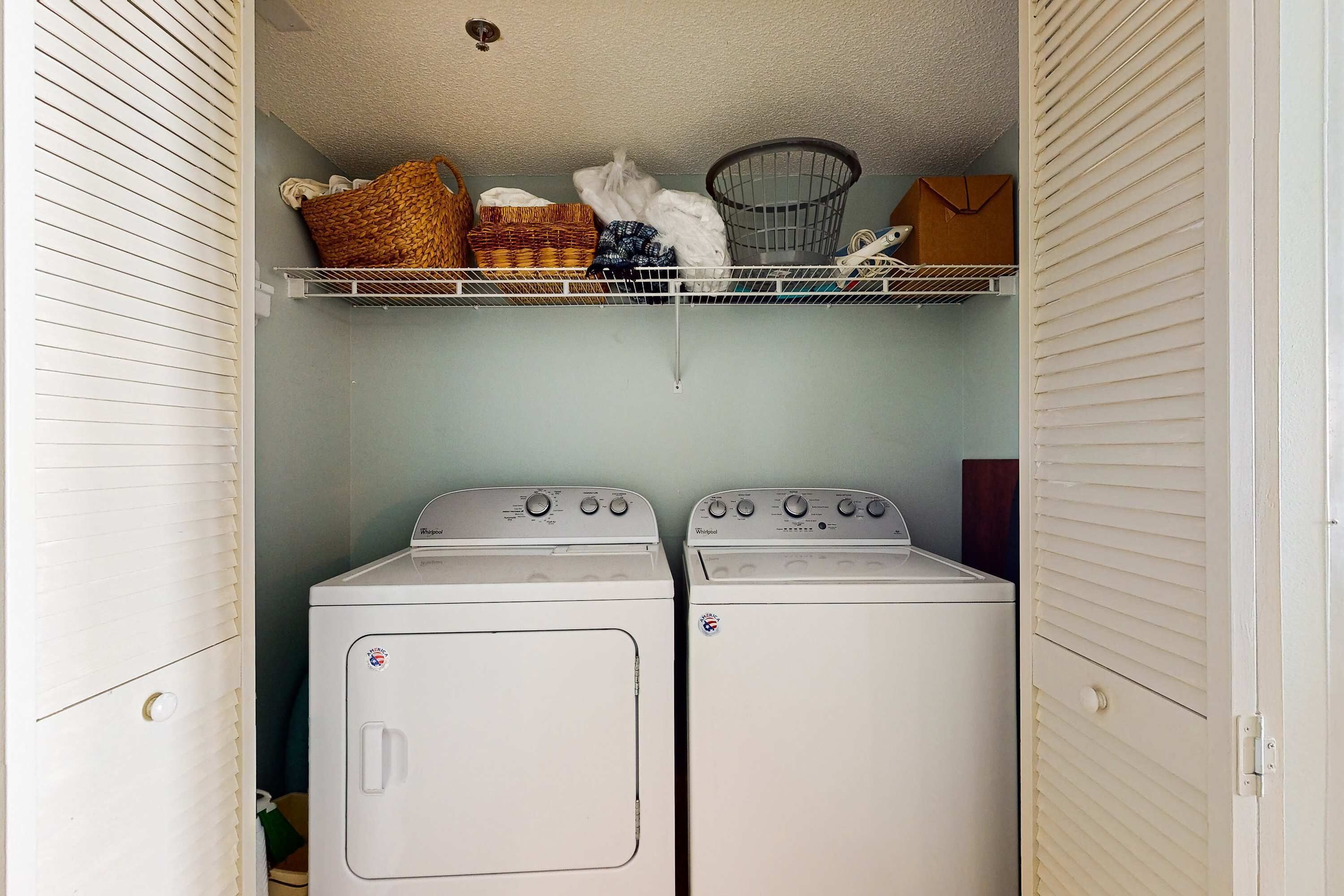 A laundry room with two stacked washers and dryers, a shelf with baskets and a basket, and a door with a handle.