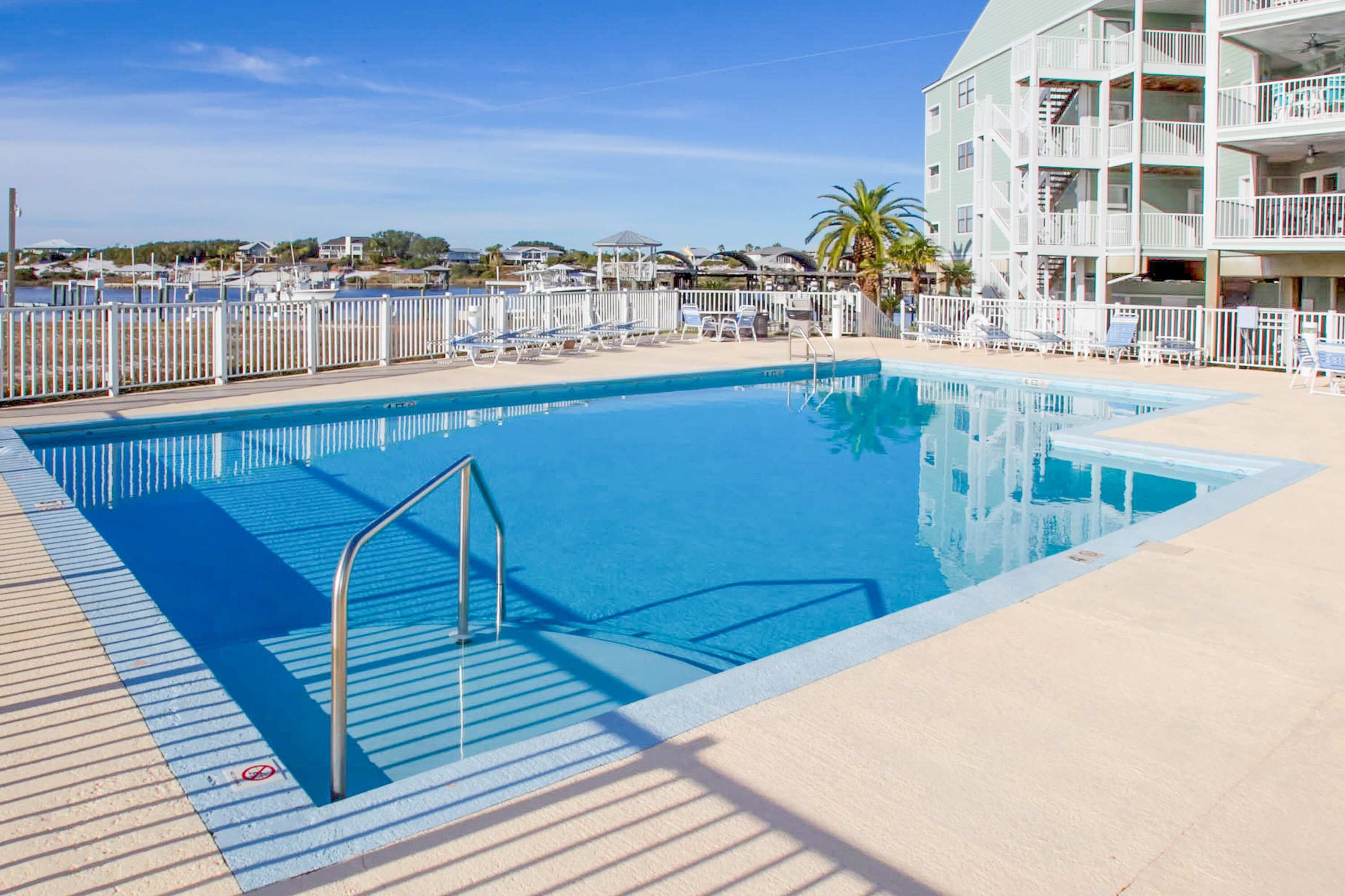 A swimming pool with a metal ladder, surrounded by a white fence, and a building with balconies in the background.