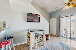 Ein Zimmer mit an der Wand montiertem Fernseher, einem kleinen Holztisch, zwei Barhocker, einem Teppich und einem Deckenventilator.
