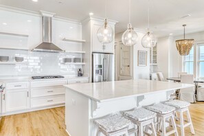 A modern kitchen with white cabinetry, a large island with bar stools, stainless steel appliances, and pendant lighting. - Luxurious Gulf-front home w private plunge pool, steps from the beach (Orange Beach)