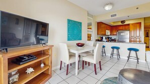 A living room with a TV, white chairs, a dining table, and a kitchen area with wooden cabinets and a refrigerator. - Cozy, beachfront condo w/ multiple shared pools, fitness center, & grilling area (Orange Beach)