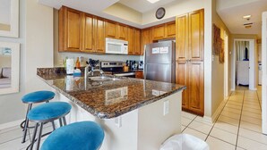 A kitchen with wooden cabinets, a granite countertop, a stainless steel refrigerator, and blue bar stools. - Cozy, beachfront condo w/ multiple shared pools, fitness center, & grilling area (Orange Beach)