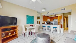 A living room with a TV, a coffee table, and a dining area with chairs and a table. - Cozy, beachfront condo w/ multiple shared pools, fitness center, & grilling area (Orange Beach)