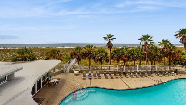 A pool area with lounge chairs, a small table, and a view of the beach. - Cozy, beachfront condo w/ multiple shared pools, fitness center, & grilling area (Orange Beach)
