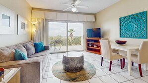 A modern living room with a sofa, round coffee table, dining table with chairs, a TV, and a balcony with a view. - Cozy, beachfront condo w/ multiple shared pools, fitness center, & grilling area (Orange Beach)