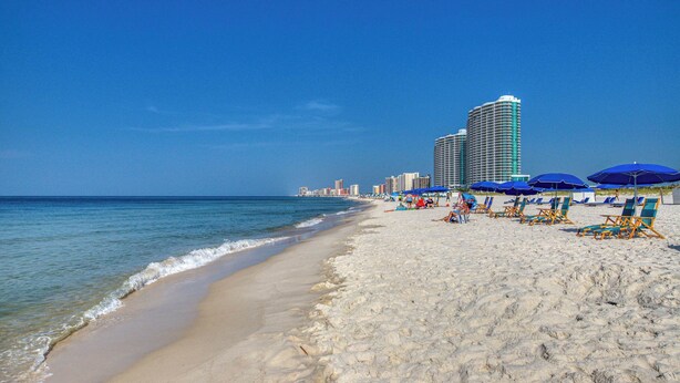 Ein Strand mit einer Reihe blauer Sonnenschirme und Liegestühlen, einem sandigen Ufer und einer Stadtansicht im Hintergrund.