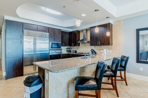 A modern kitchen with dark wood cabinets, stainless steel appliances, a granite island, and bar stools. - Gulf-front condo with pool, tennis, lazy river & private hot tub (Orange Beach)