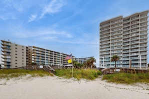 Ein Strandhotel mit mehreren Balkonen, einem Sandstrand und einer Fahnenmast mit Flaggen.