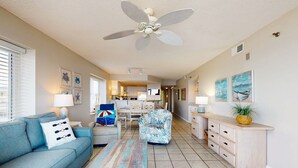 A living room with a sofa, coffee table, and a ceiling fan. There is a kitchen area in the background. - Beachfront condo w/ private, corner balcony, community pools, & a grilling area (Orange Beach)