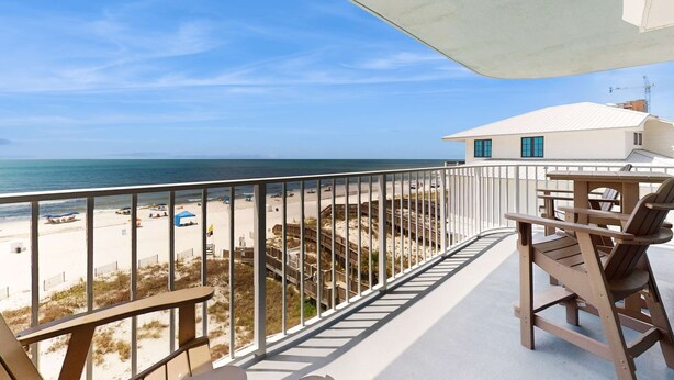 A balcony with a view of the beach, a wooden table and chairs, and a white building in the background. - Beachfront condo w/ private, corner balcony, community pools, & a grilling area (Orange Beach)