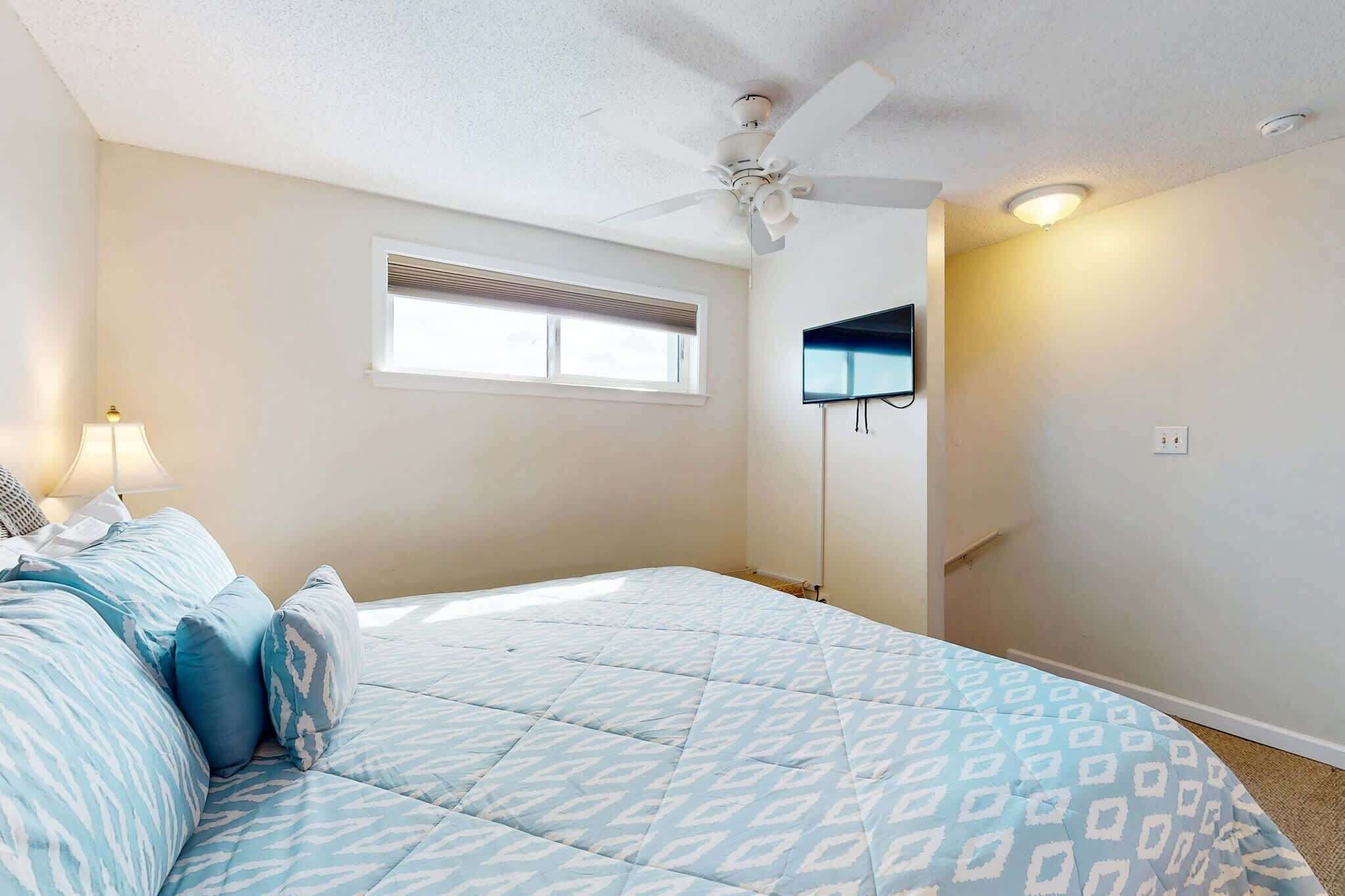 Un dormitorio con cama, ventilador de techo, ventana y televisor montado en la pared.