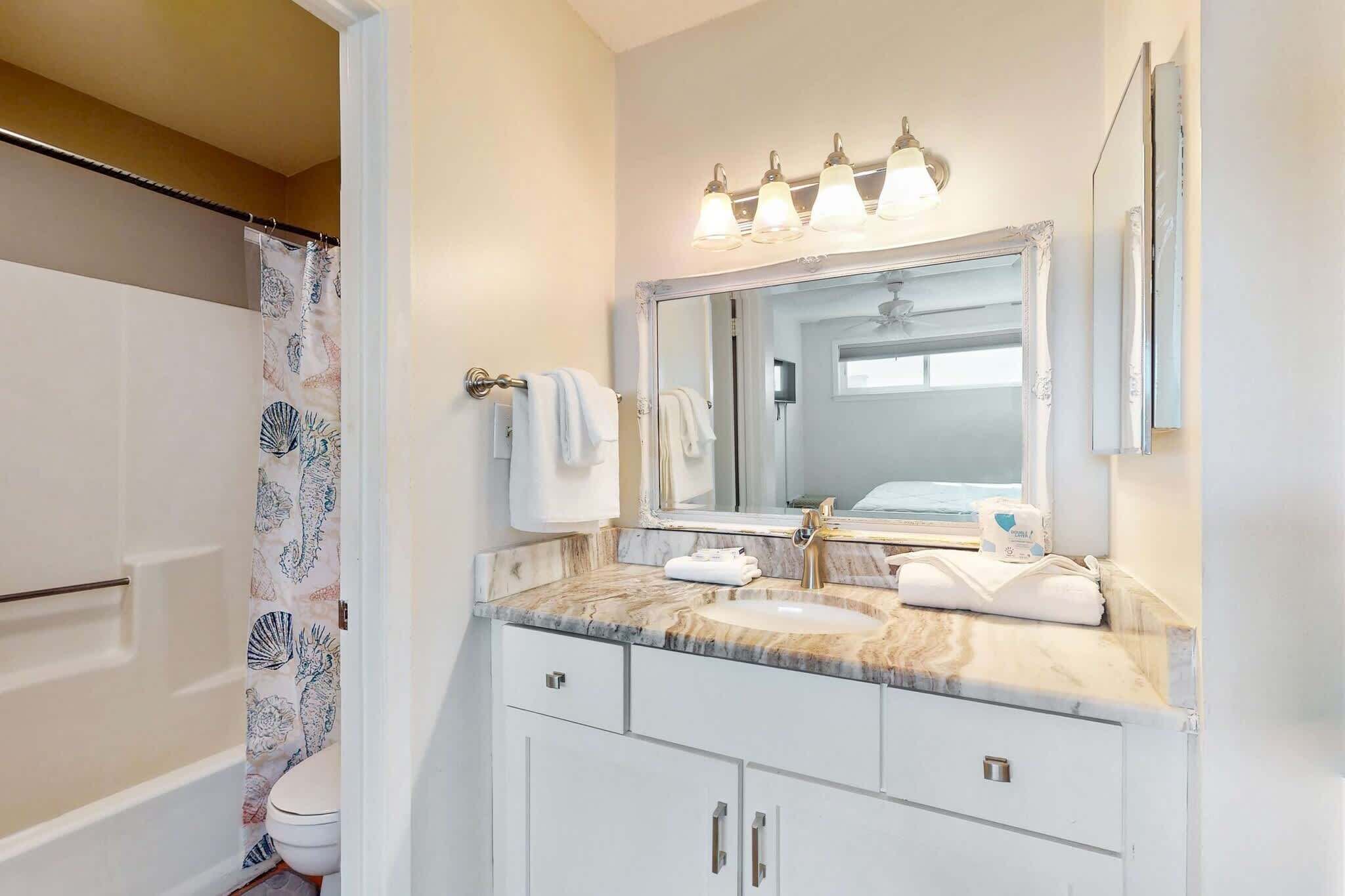 Une salle de bain avec un rideau de douche, un lavabo doté d’un plan de travail en marbre, un miroir avec éclairage intégré et un porte-serviettes.
