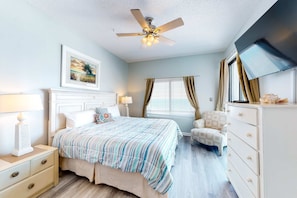 A bedroom with a bed, a ceiling fan, a window with curtains, a chair, a dresser, and a TV. - Gulf front condo with pools/hot tub - ocean view, covered balcony & washer/dryer (Orange Beach)