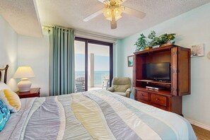 A hotel room with a bed, a television on a wooden cabinet, a ceiling fan, a window with curtains, and a view of the outdoors. - Gulf front condo w/ a private, furnished balcony, shared pools, hot tub, gym (Orange Beach)