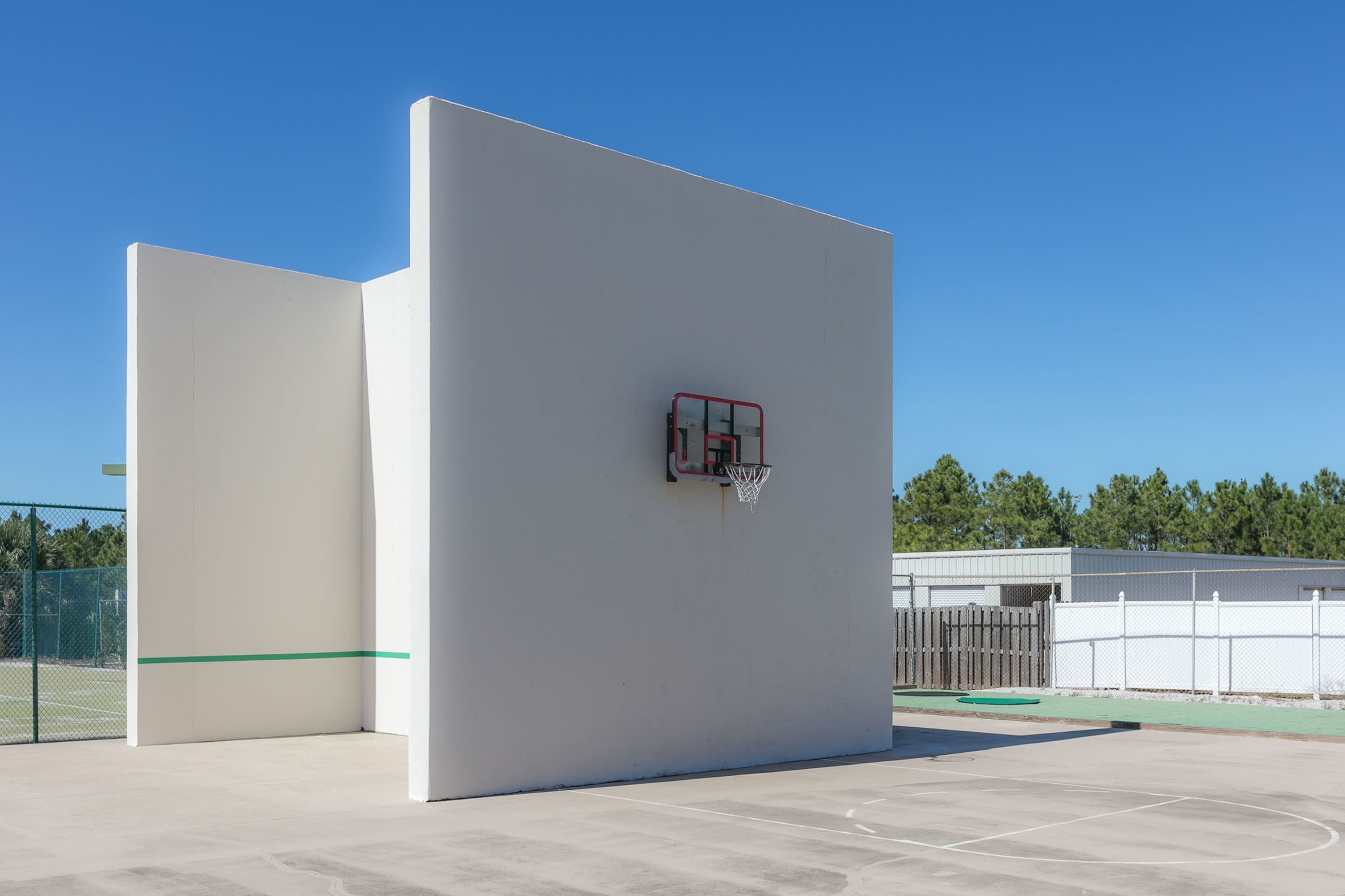 Ein Basketballplatz mit Korb, Zaun und Bäumen im Hintergrund.