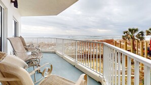 Een balkon met rieten stoelen, een witte balustrade en uitzicht op het strand en de zee.