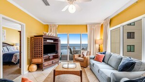 A living room with a sofa, coffee table, TV stand, and a balcony with a view. - Beachfront condo with Gulf front shared pool, private balcony, and grilling area (Orange Beach)