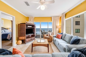A living room with a sofa, coffee table, TV stand, and a balcony with a view. - Beachfront condo with Gulf front shared pool, private balcony, and grilling area (Orange Beach)