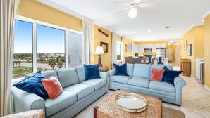 A spacious living area with two light blue sofas, a woven coffee table, and a view of the outdoors through large windows. - Beachfront condo with Gulf front shared pool, private balcony, and grilling area (Orange Beach)
