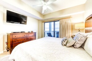 A hotel room with a bed, a TV mounted on the wall, a ceiling fan, a dresser, and a view of the outdoors through a large window. - Beachfront condo with shared outdoor pool, heated indoor pool, sauna, & grill area (Orange Beach)