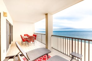 A balcony with a table and chairs, a small stool, and a view of the ocean. - Beachfront condo with shared outdoor pool, heated indoor pool, sauna, & grill area (Orange Beach)