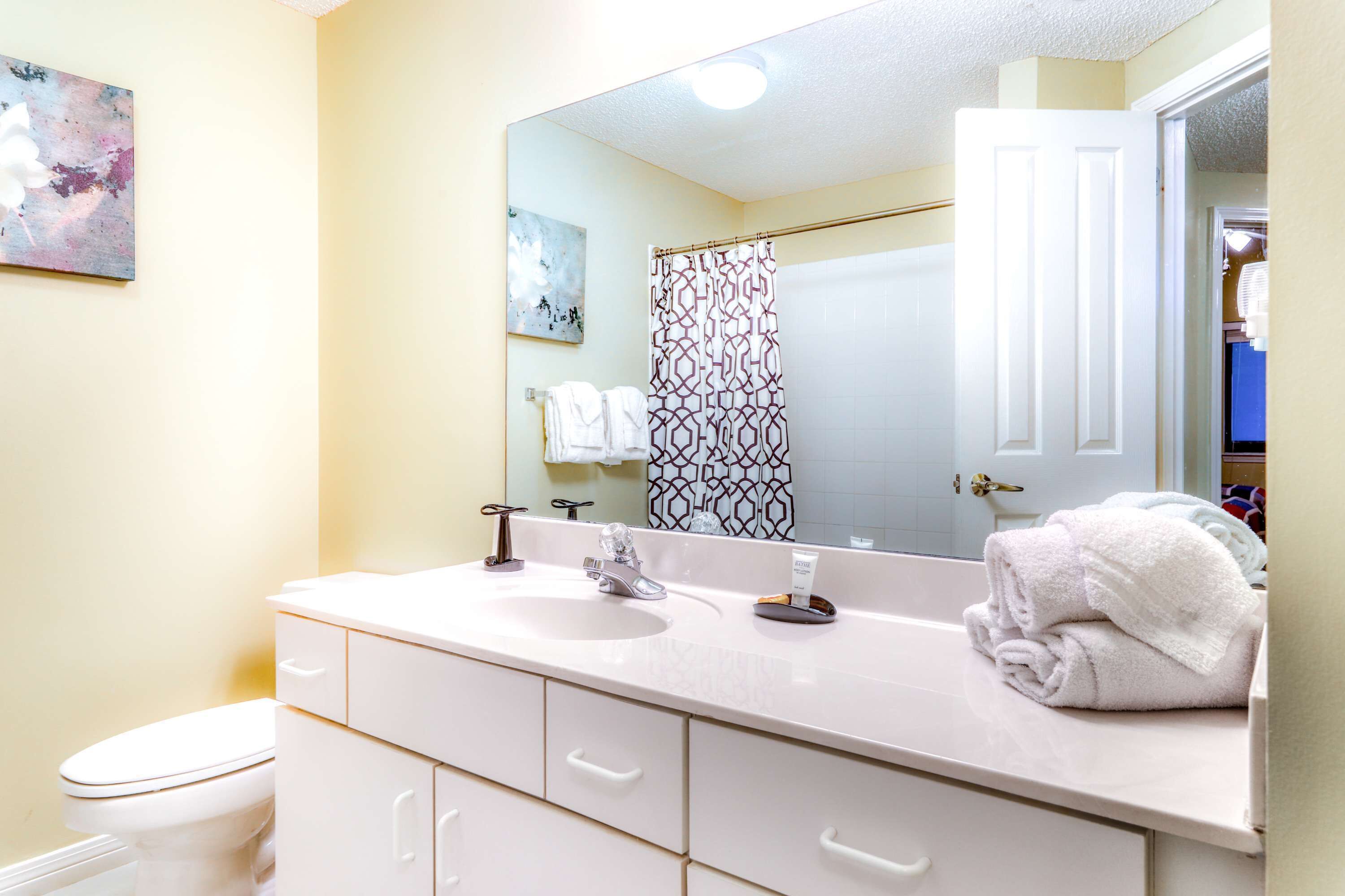 Une salle de bain avec un lavabo blanc, deux robinets, un miroir, un rideau de douche et des toilettes.