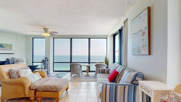 A spacious living room with a large window, a ceiling fan, and a view of the ocean. - Gulf-front condo with deep corner balcony, beach view, pools & gym (Orange Beach)