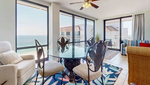 A room with a glass table surrounded by chairs, a ceiling fan, large windows with a view of the ocean, and a balcony with a view. - Gulf-front condo with deep corner balcony, beach view, pools & gym (Orange Beach)