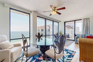 A room with a glass table surrounded by chairs, a ceiling fan, large windows with a view of the ocean, and a balcony with a view. - Gulf-front condo with deep corner balcony, beach view, pools & gym (Orange Beach)