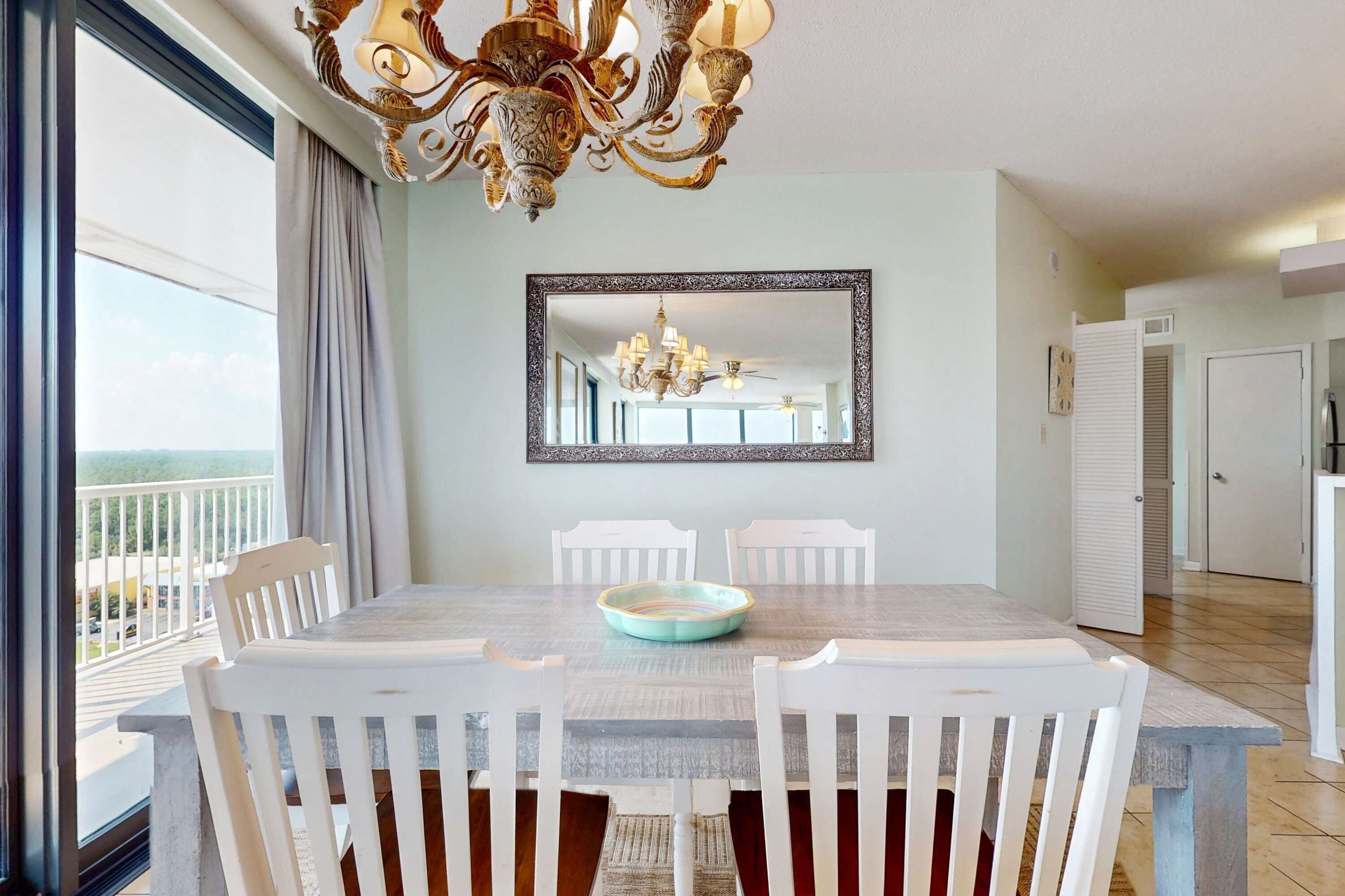 A dining area with a large table, white chairs, a chandelier, and a view through a balcony.