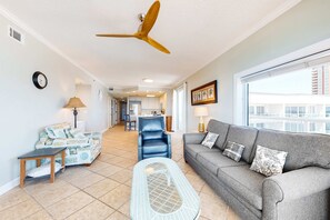 Ein Wohnzimmer mit Deckenventilator, einer Couch, einem Sessel, einem kleinen Tisch sowie Blick auf Küche und Balkon.